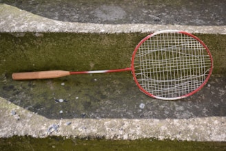 A worn badminton racket with a red and white frame is resting on a moss-covered concrete step. The racket's strings are slightly sagging, indicating it is old or well-used. The surrounding surface has patches of green and gray, with some small stones and worn areas visible.