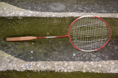 A worn badminton racket with a red and white frame is resting on a moss-covered concrete step. The racket's strings are slightly sagging, indicating it is old or well-used. The surrounding surface has patches of green and gray, with some small stones and worn areas visible.