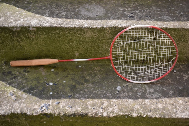 A worn badminton racket with a red and white frame is resting on a moss-covered concrete step. The racket's strings are slightly sagging, indicating it is old or well-used. The surrounding surface has patches of green and gray, with some small stones and worn areas visible.