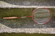 A worn badminton racket with a red and white frame is resting on a moss-covered concrete step. The racket's strings are slightly sagging, indicating it is old or well-used. The surrounding surface has patches of green and gray, with some small stones and worn areas visible.