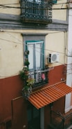 An exterior view of a building showcasing a window with ornate wrought iron grilles. Below the window is a small balcony adorned with various potted plants, adding a touch of greenery. An air conditioning unit is mounted on the wall beside the balcony. The building has a two-tone color scheme, with a yellow upper facade and a red-brown lower section. A terracotta-colored awning covers the entrance below, hinting at Mediterranean architectural influences.