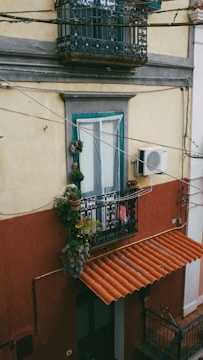 An exterior view of a building showcasing a window with ornate wrought iron grilles. Below the window is a small balcony adorned with various potted plants, adding a touch of greenery. An air conditioning unit is mounted on the wall beside the balcony. The building has a two-tone color scheme, with a yellow upper facade and a red-brown lower section. A terracotta-colored awning covers the entrance below, hinting at Mediterranean architectural influences.