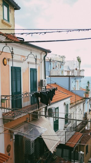 A Mediterranean-style street scene featuring buildings with light peach and white facades, green shutters, and terracotta roof tiles. Clothes are hung to dry on a balcony railing, and a narrow street is visible below.