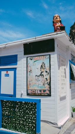 A colorful kiosk with blue and white wooden elements features a large poster with a retro aesthetic. On top, a sculpture of a person's head adds a whimsical touch. The kiosk has a door and a small green fence made of artificial foliage. A sign next to the door indicates a toilet. A quote is displayed on a board on the side of the building. The sky is clear with a few clouds.