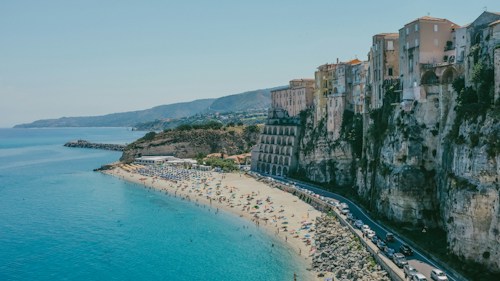A picturesque coastal scene featuring a sandy beach lined with colorful umbrellas and numerous people enjoying the seaside. To the right, there is a dramatic cliffside with a series of historic buildings perched on top, showcasing a blend of architectural styles. The clear blue water of the ocean contrasts beautifully with the beige of the sand and the white and pastel colors of the buildings. A winding road runs below the cliff, with vehicles traveling along it. The backdrop features gently rolling hills under a clear blue sky.