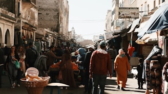 A bustling street market with various stalls and vendors selling goods. People are walking, shopping, and engaging with the vendors. The buildings lining the street have an old architectural style, and there are signs in a language that appears to be Arabic. The atmosphere seems lively with a mix of colorful items on display.
