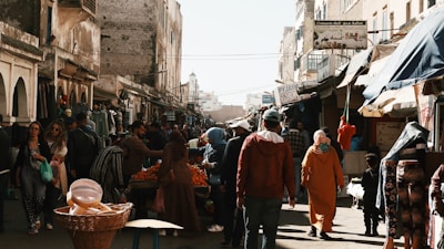 A bustling street market with various stalls and vendors selling goods. People are walking, shopping, and engaging with the vendors. The buildings lining the street have an old architectural style, and there are signs in a language that appears to be Arabic. The atmosphere seems lively with a mix of colorful items on display.