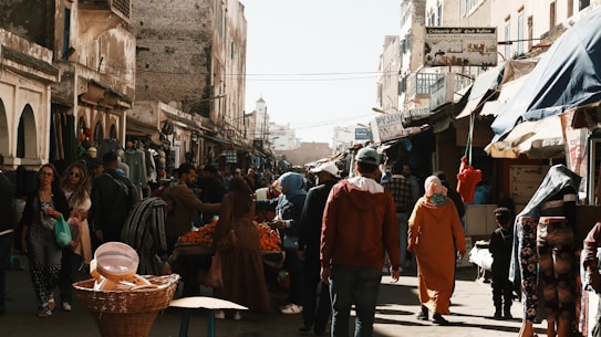 A bustling street market with various stalls and vendors selling goods. People are walking, shopping, and engaging with the vendors. The buildings lining the street have an old architectural style, and there are signs in a language that appears to be Arabic. The atmosphere seems lively with a mix of colorful items on display.