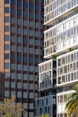 Two contrasting building facades appear side by side. On the left, a brown brick high-rise features a repetitive pattern of dark, rectangular windows. On the right, a modern, white building is characterized by large, protruding balconies and extensive use of glass, with visible greenery on some balconies. In the foreground, the tops of trees are visible, including a palm tree.
