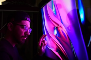 A person with glasses and a beard is wearing a black hat and is closely observing or interacting with a digitally created colorful abstract image on a screen. The abstract design features swirling patterns of bright pink, blue, and purple hues.