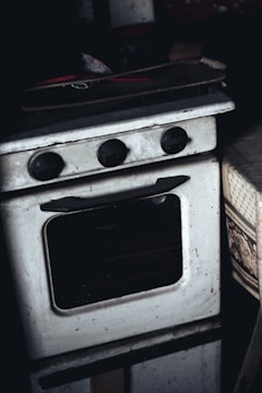 A vintage, slightly grimy white oven with black control knobs is placed in a dimly lit environment. The oven appears worn, with visible signs of use and some dirt. A wooden tray or board with red-handled utensils or tools rests on top of the oven. To the right, part of a patterned fabric, possibly a tablecloth, is visible.