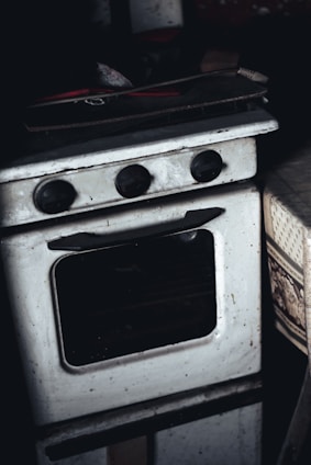 A vintage, slightly grimy white oven with black control knobs is placed in a dimly lit environment. The oven appears worn, with visible signs of use and some dirt. A wooden tray or board with red-handled utensils or tools rests on top of the oven. To the right, part of a patterned fabric, possibly a tablecloth, is visible.