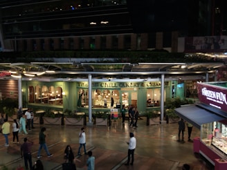 An outdoor view of a bakery and creamery with a green facade, visible through large windows. The establishment has warm interior lighting, and several people are walking or standing in the pedestrian area outside. A kiosk selling frozen treats is nearby, with vibrant pink and purple signage. The scene occurs at night, with ambient lighting highlighting the shop windows and the walkway.
