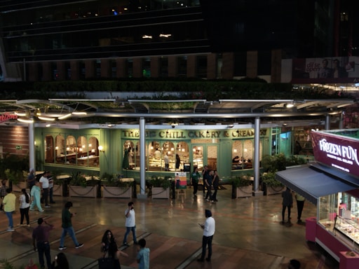 An outdoor view of a bakery and creamery with a green facade, visible through large windows. The establishment has warm interior lighting, and several people are walking or standing in the pedestrian area outside. A kiosk selling frozen treats is nearby, with vibrant pink and purple signage. The scene occurs at night, with ambient lighting highlighting the shop windows and the walkway.