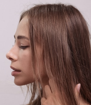 A profile view of a person with long brown hair. The individual's eyes are closed, and they appear serene. A hand gently touches the neck, suggesting a contemplative or relaxed state. The background is plain and muted, drawing focus to the subject.