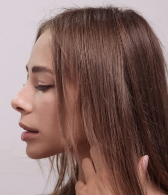 A profile view of a person with long brown hair. The individual's eyes are closed, and they appear serene. A hand gently touches the neck, suggesting a contemplative or relaxed state. The background is plain and muted, drawing focus to the subject.