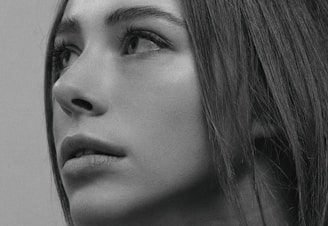A close-up black and white portrait of a person gazing upward, capturing intricate details of the facial features, including the eyes, eyelashes, nose, and lips. The person's hair is visible, framing the face and adding depth to the image.