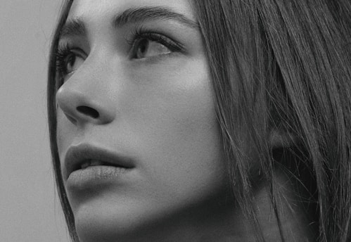 A close-up black and white portrait of a person gazing upward, capturing intricate details of the facial features, including the eyes, eyelashes, nose, and lips. The person's hair is visible, framing the face and adding depth to the image.