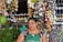 A woman smiles while standing in front of a stall lined with an assortment of colorful bottles and herbs. Various liquids in small containers hang around her, creating a vibrant display. She wears a patterned blouse and a green apron, surrounded by an array of products, suggesting a market setting.