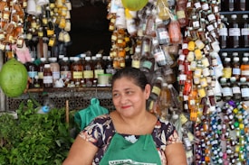 A woman smiles while standing in front of a stall lined with an assortment of colorful bottles and herbs. Various liquids in small containers hang around her, creating a vibrant display. She wears a patterned blouse and a green apron, surrounded by an array of products, suggesting a market setting.