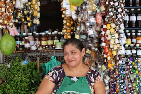 A woman smiles while standing in front of a stall lined with an assortment of colorful bottles and herbs. Various liquids in small containers hang around her, creating a vibrant display. She wears a patterned blouse and a green apron, surrounded by an array of products, suggesting a market setting.