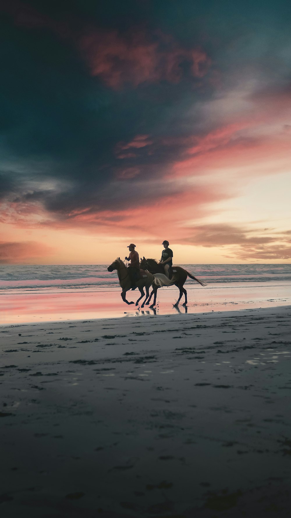 two people riding horses on a beach at sunset