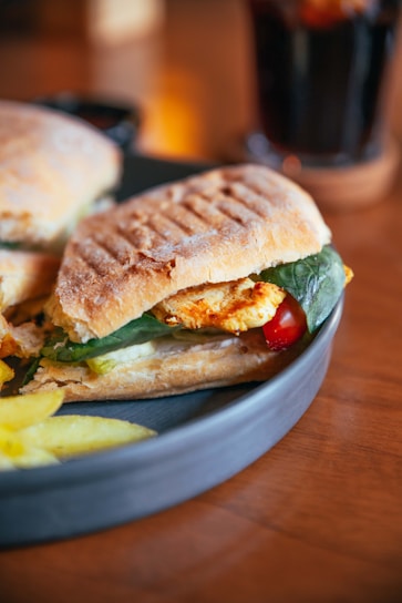 Two grilled sandwiches with crispy bread are placed on a gray plate. The sandwiches contain spinach, slices of red bell pepper, chicken, and cheese. There are a few potato chips on the plate. The background is slightly blurred, showing a brown surface with a dark beverage in a glass.