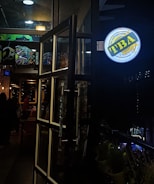 A dimly lit entrance to a bar or cafe with open glass doors framed in metal. The interior is visible, showing some people inside, a wall adorned with colorful graffiti, and ceiling lights that contribute to a cozy ambiance. A round, illuminated sign hangs outside with writing in multiple languages.