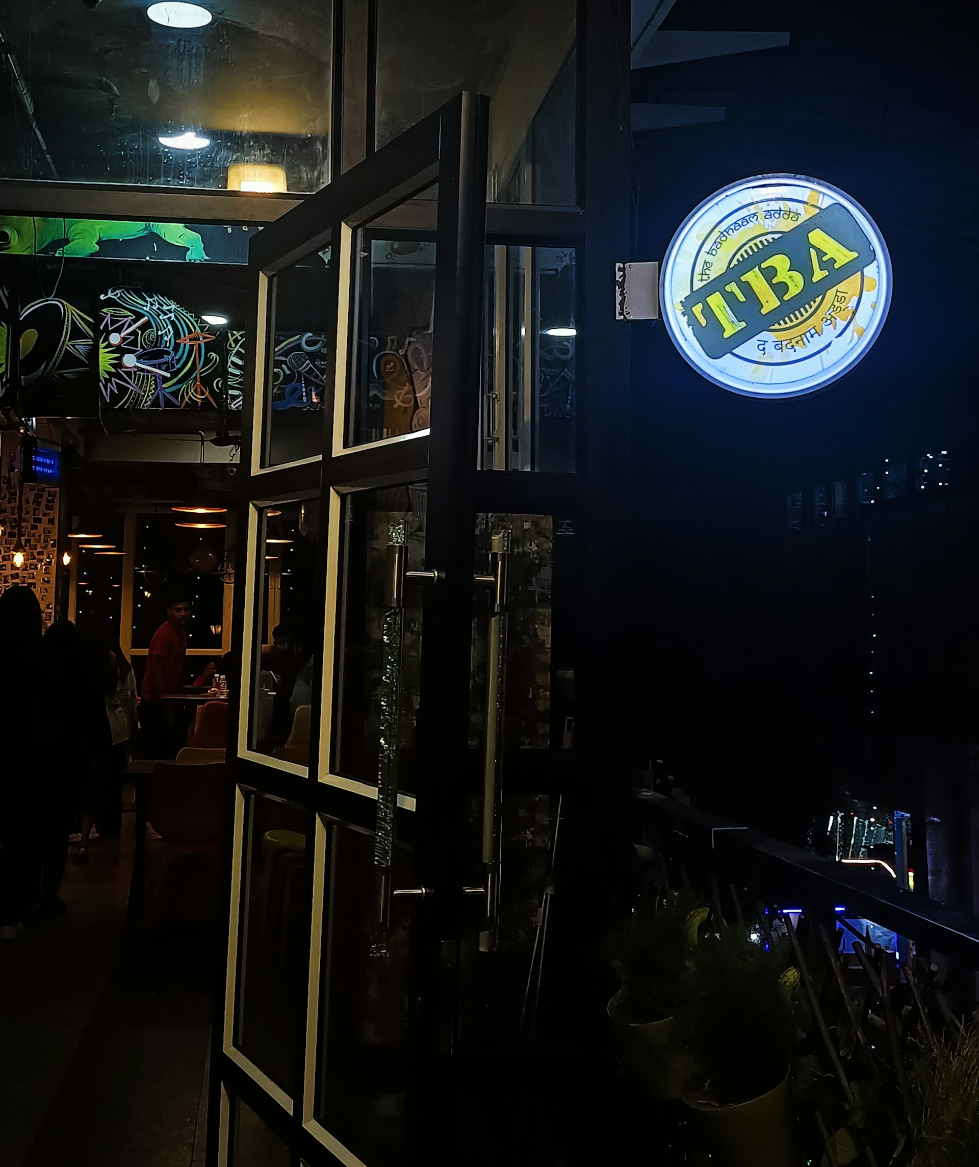 A dimly lit entrance to a bar or cafe with open glass doors framed in metal. The interior is visible, showing some people inside, a wall adorned with colorful graffiti, and ceiling lights that contribute to a cozy ambiance. A round, illuminated sign hangs outside with writing in multiple languages.