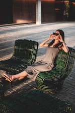 A woman is lounging comfortably on a set of dark green outdoor chairs, wearing sunglasses and a casual gray dress. The setting is outdoors, with a paved ground and shaded, ambient lighting creating a relaxed atmosphere. The woman is stretching out her legs, appearing at ease, as she enjoys the serene environment.
