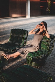 A woman is lounging comfortably on a set of dark green outdoor chairs, wearing sunglasses and a casual gray dress. The setting is outdoors, with a paved ground and shaded, ambient lighting creating a relaxed atmosphere. The woman is stretching out her legs, appearing at ease, as she enjoys the serene environment.