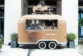 A tan food truck with a retro design is parked between two large planters. The truck features a service window displaying bottles and condiments. A person inside is partially visible through the window.