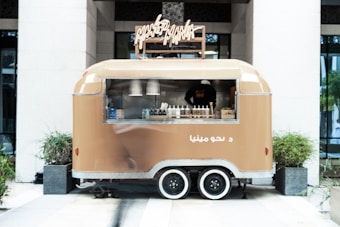 A tan food truck with a retro design is parked between two large planters. The truck features a service window displaying bottles and condiments. A person inside is partially visible through the window.