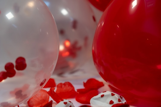 Red and translucent balloons filled with small red objects are surrounded by scattered red rose petals. The atmosphere is festive, with a focus on the contrasting textures and colors. A dessert topped with small red heart-shaped sprinkles is also visible among the decorations.