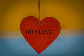 A red heart-shaped pendant hangs by a string against a backdrop that blends yellow and blue. The words 'WITH LOVE' are embossed in the center of the heart.