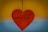 A red heart-shaped pendant hangs by a string against a backdrop that blends yellow and blue. The words 'WITH LOVE' are embossed in the center of the heart.