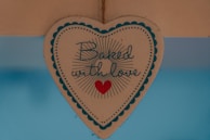 A heart-shaped sign with scalloped edges and a decorative border contains the phrase 'Baked with love' written in a cursive font, accompanied by a small red heart icon. The sign is suspended against a blue background.
