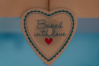 A heart-shaped sign with scalloped edges and a decorative border contains the phrase 'Baked with love' written in a cursive font, accompanied by a small red heart icon. The sign is suspended against a blue background.