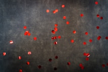 A dark textured background scattered with numerous red circular confetti pieces. The red circles are bright and reflective, creating a contrast against the subdued background.