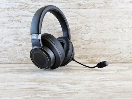 A black gaming headset with attached microphone is displayed on a light wooden surface. The background also features a wooden texture in the same light tone.