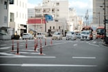 An urban street scene with multiple cars and buses stopped at an intersection. Several orange traffic cones are aligned on the road, guiding vehicles. Surrounding buildings include multi-story structures and storefronts with visible signage, and overhead power lines crisscross above, reflecting a typical city environment.