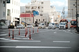 An urban street scene with multiple cars and buses stopped at an intersection. Several orange traffic cones are aligned on the road, guiding vehicles. Surrounding buildings include multi-story structures and storefronts with visible signage, and overhead power lines crisscross above, reflecting a typical city environment.