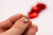A hand holding a gold ring with three embedded diamonds, set against a blurred red background which appears to be a shape, possibly a ribbon or decoration.