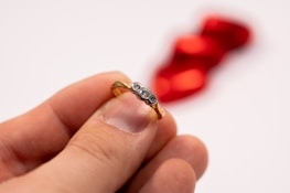 A hand holding a gold ring with three embedded diamonds, set against a blurred red background which appears to be a shape, possibly a ribbon or decoration.