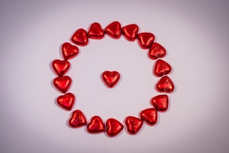 A collection of red, heart-shaped objects arranged in a circle on a light-colored surface, with one heart positioned in the center of the circle.