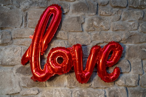 Red foil balloon letters spelling the word 'love' are mounted against a rustic stone wall.