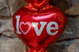 A shiny, red heart-shaped balloon with the word 'love' printed on it in bold white letters. The background consists of gray stones, giving a rustic feel.