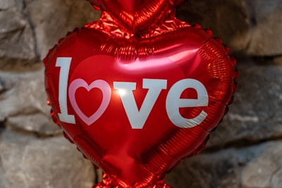 A shiny, red heart-shaped balloon with the word 'love' printed on it in bold white letters. The background consists of gray stones, giving a rustic feel.