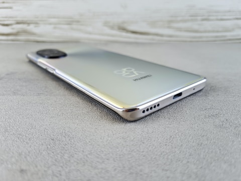 A sleek, modern smartphone with a reflective back surface rests on a textured surface. The branding and logo are visible, along with the camera module at the top left. The phone's edges are smooth and metallic, and the bottom features a USB port and speaker holes.