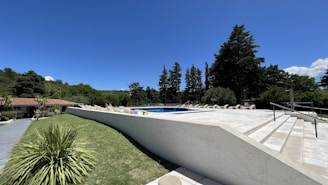 A serene outdoor setting featuring a large, tiled pool area with several lounge chairs neatly aligned along the pool. The surrounding landscape is lush with green grass and tall trees. A small building with a red-tiled roof is visible to the left, and the sky is clear with deep blue colors.